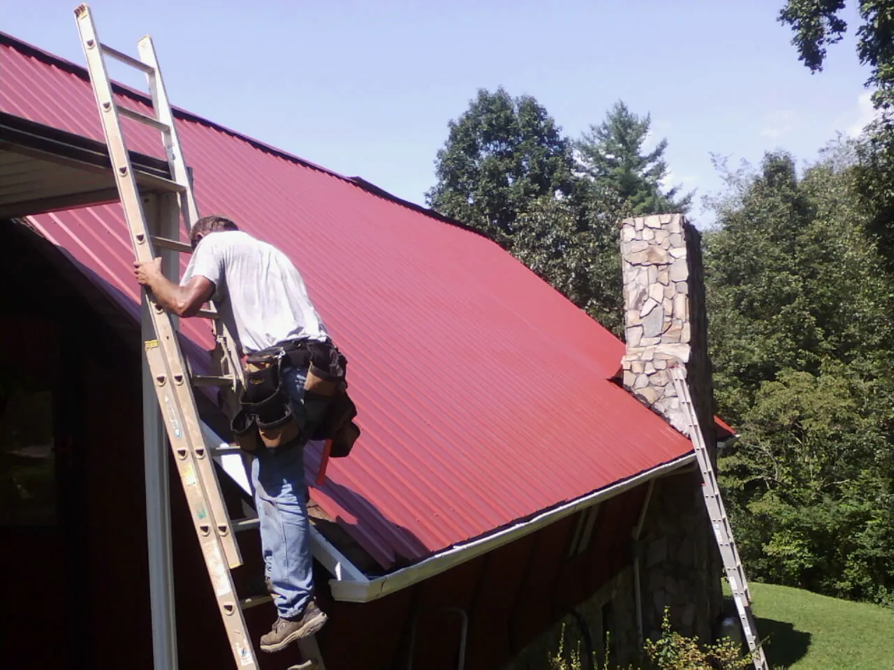 Roofer on a ladder beside a red metal roof and stone chimney — Soffit & Fascia Repair by Roofing Brothers in Robinson